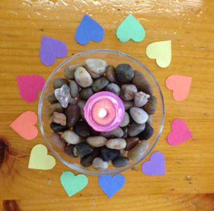 Photograph of a single lit candle surrounded by colorful paper hearts.